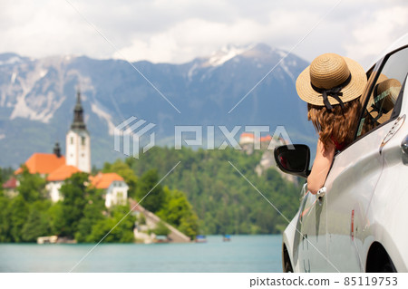 Happy young woman on vacation leaning out of car window on shore of Bled lake, country of Slovenia. Travel, holiday, tourism, explore, rent a car concept. Happy young woman on vacation leaning out of car window on shore of Bled lake, country of Slovenia. Travel, holiday, tourism, explore, rent a car concept. 85119753