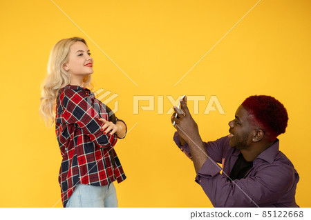 Selfie making. Social media. Mobile technology. Multiracial relationship. Diverse couple man taking photo on phone for blog of posing woman isolated on yellow background. Selfie making. Social media. Mobile technology. Multiracial relationship. Diverse couple man taking photo on phone for blog of posing woman isolated on yellow background. 85122668