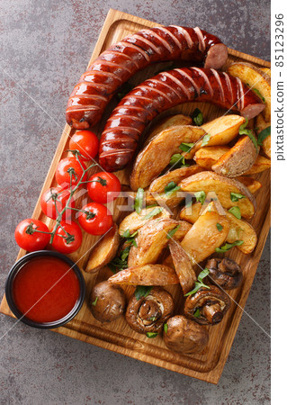 Grilled sausages with potato wedges and mushrooms close-up on a wooden tray. vertical top view Grilled sausages with potato wedges and mushrooms close-up on a wooden tray. vertical top view 85123296