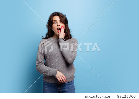 Studio shot of shocked young pretty girl in warm gray knitted sweater posing isolated on blue color background Studio shot of shocked young pretty girl in warm gray knitted sweater posing isolated on blue color background 85126306