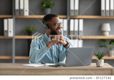 Satisfied millennial black bearded man in glasses and headphones works at laptop and looks at empty space Satisfied millennial black bearded man in glasses and headphones works at laptop and looks at empty space 85127192