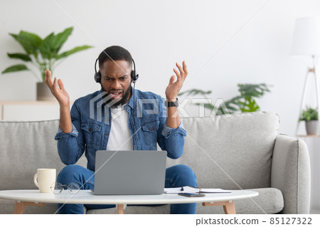Shocked displeased young black bearded man in headphones raises hands, talk by laptop and gesturing 85127322