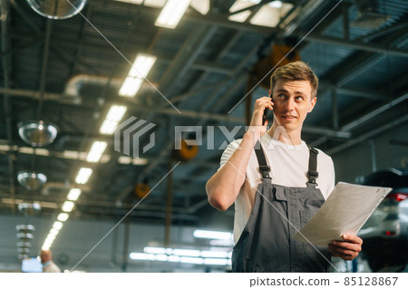 Low-angle view of professional handsome young mechanic male wearing uniform holding clipboard and talking on mobile phone. Low-angle view of professional handsome young mechanic male wearing uniform holding clipboard and talking on mobile phone. 85128867