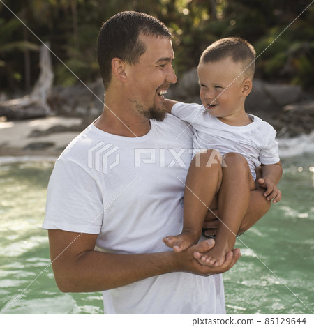 A black-haired young bearded father holds in arms his little child with a blond son 85129644