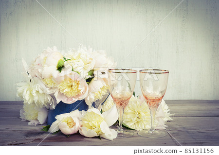 Beautiful celebration composition: Two beautiful empty wineglass, bouquet of white peonies on rustic wooden background; copy space 85131156
