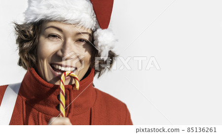 Curly winking woman in Santa Claus hat bites striped candy cane. Pretty female with Christmas licorice whip. Banner with copy space. 85136261