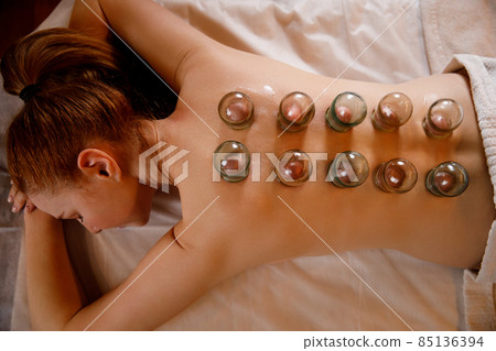 A young woman relaxes in a massage parlor. Vacuum cups of medical cupping therapy on woman back, close up, chinese medicine. A young woman relaxes in a massage parlor. Vacuum cups of medical cupping therapy on woman back, close up, chinese medicine. 85136394