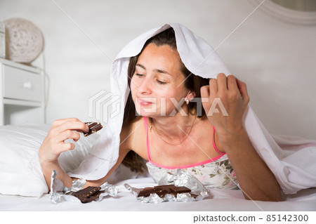 Cute young woman in sleepwear eating chocolate in bed at home Cute young woman in sleepwear eating chocolate in bed at home 85142300