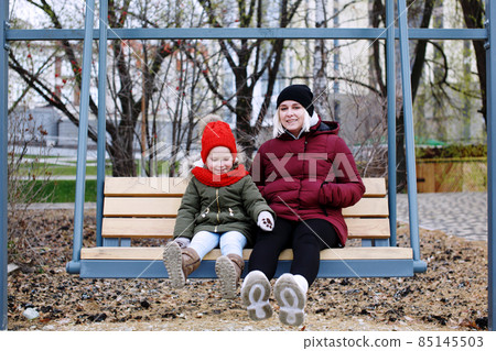Young mother talking to her cute little daughter outdoors sitting together in warm clothes Young mother talking to her cute little daughter outdoors sitting together in warm clothes 85145503