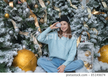 New Year portrait of beautiful caucasian young woman in cozy light blue sweater and hat. Adorable smiling girl sitting near decorated Christmas tree 85149874