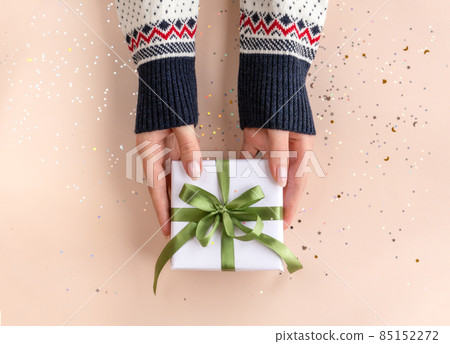 Female hands in sweater with pink manicure holding present in white paper with green ribbon on soft beige background. Xmas composition. Flat lay. Happy holidays, New Year celebration concept. Female hands in sweater with pink manicure holding present in white paper with green ribbon on soft beige background. Xmas composition. Flat lay. Happy holidays, New Year celebration concept. 85152272