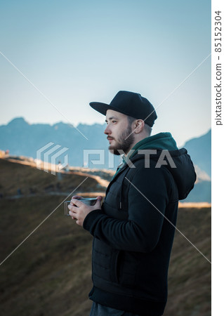 Bearded traveler man drinks tea or coffee from  metal cup while resting on a rock 85152304
