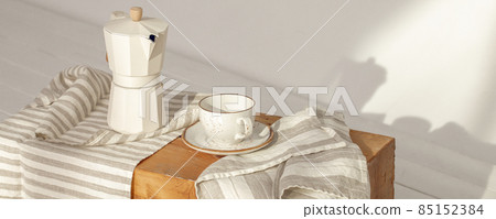 Italian coffee maker with wooden handle and white coffee craft cup on solid wood stamp decorated with tea towels in creamy tones on sunlight creating beautiful shadows on the wall Italian coffee maker with wooden handle and white coffee craft cup on solid wood stamp decorated with tea towels in creamy tones on sunlight creating beautiful shadows on the wall 85152384