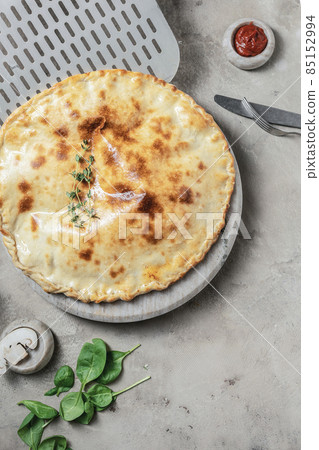 Italian calzone pizza with mozzarella, ham, basil on marble round tray. Garnished on grey stone backdrop with pizza ingredients Italian calzone pizza with mozzarella, ham, basil on marble round tray. Garnished on grey stone backdrop with pizza ingredients 85152994