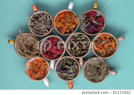 Assortment of dried relaxing tea herbs in colourful cups on blue background close up. Calendula, mint, anise hyssop, monarda didyma, wormwood, sage leaves. Assortment of dried relaxing tea herbs in colourful cups on blue background close up. Calendula, mint, anise hyssop, monarda didyma, wormwood, sage leaves. 85153002