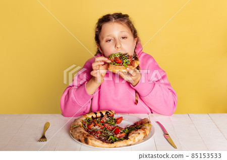 Studio shot of little cute girl sitting and tasting delicious Italian pizza isolated on yellow studio background. 85153533