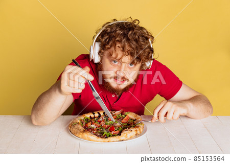 Cunning red-bearded man cutting and eating delicious pizza with cheese and tomatoes isolated on yellow studio background. 85153564