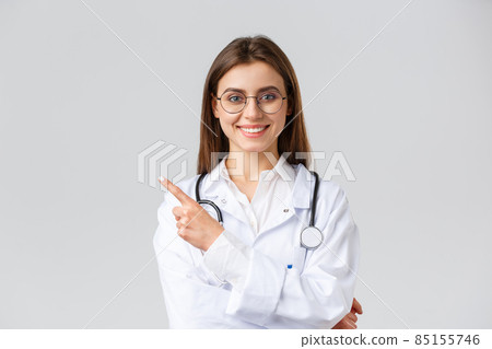 Healthcare workers, medicine and covid-19 pandemic concept. Good-looking professional female doctor in white scrubs and glasses, pointing fingers left, smiling friendly, showing information 85155746