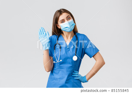 Covid-19, preventing virus, health, healthcare workers and quarantine concept. Friendly female nurse in clinic wearing blue scrubs and medical mask, waving hello or hi sign with gloves on 85155845