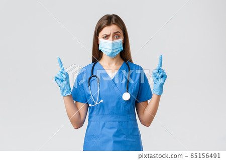 Covid-19, preventing virus, health, healthcare workers and quarantine concept. Skeptical female nurse in blue scrubs and medical mask dont believe this info, pointing fingers up doubtful 85156491