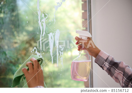 Close-up of the hands of a woman cleansing the window, removing stains by spraying cleaning products and wiping with a sponge. Spring house cleaning concept with copy ad space 85156712