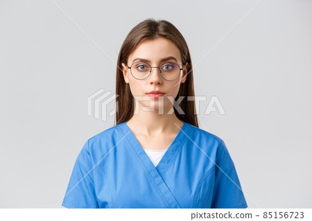 Healthcare workers, medicine, insurance and covid-19 pandemic concept. Young female intern, nurse or doctor in scrubs and glasses, looking at camera with serious expression, grey background 85156723