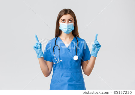 Covid-19, preventing virus, health, healthcare workers and quarantine concept. Curious female doctor or nurse in blue scrubs, medical mask reading banner, pointing fingers up 85157230