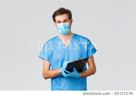 Covid-19, quarantine, hospitals and healthcare workers concept. Smiling young doctor, nurse in medical mask, scrubs and gloves, writing down patient symptoms or info at clipboard, fill in blank in ER 85157276
