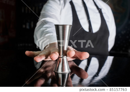 The hand of a professional bartender holds a jigger tool or measuring cup to control the ingredients added to the cocktail 85157763