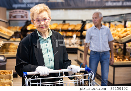 an elderly woman with glasses is shopping in the bread department of a supermarket 85159192