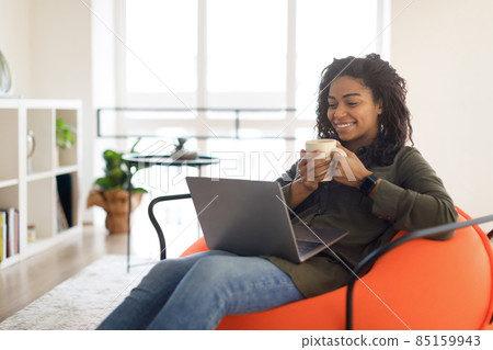 Smiling black woman watching video on computer, drinking coffee Smiling black woman watching video on computer, drinking coffee 85159943
