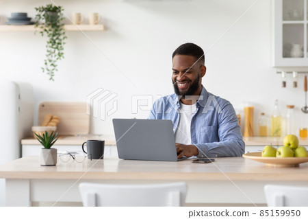 Glad millennial african american bearded male working on laptop in scandinavian kitchen interior, free space Glad millennial african american bearded male working on laptop in scandinavian kitchen interior, free space 85159950