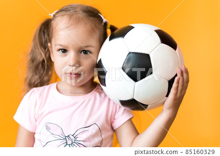 Authentic cute smiling preschool little girl with classic black and white soccer ball look at camera on yellow background. child play football in t-shirt and shorts. Sport, championship, team concept 85160329