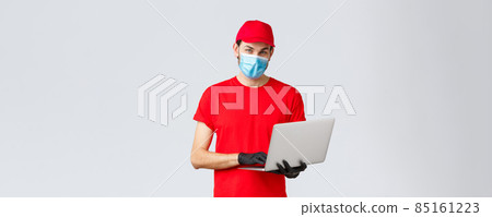 Customer support, covid-19 delivery packages, online orders processing concept. Pleasant delivery guy in red uniform, face mask and gloves, using laptop and look camera, processing order 85161223
