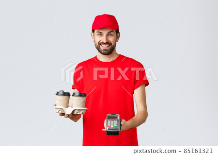 Food delivery, quarantine, stay home and order online concept. Friendly bearded courier in red uniform handing coffee delivery and POS terminal for client to pay contactless, grey background 85161321