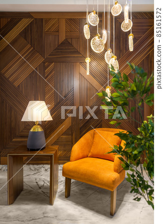 Modern orange armchair, lamp shade on small wooden table, contemporary tall glass chandelier, and wood cladding wall. 85161572
