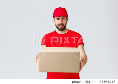 Order delivery, online shopping and package shipping concept. Cheeky handsome bearded courier in red uniform, handing box package to client. Employee wink to you and give order parcel 85161937