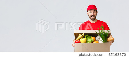 Groceries and packages delivery, covid-19, quarantine and shopping concept. Smiling handsome bearded courier in red uniform, bring food package, grocery order to client in box, look amused 85162870