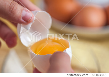 Woman hands breaking an egg to separate egg white and yolks, egg shells at the background Woman hands breaking an egg to separate egg white and yolks, egg shells at the background 85163368