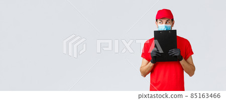 Packages, delivery, covid-19 quarantine, transfer orders. Curious and excited courier in red uniform, gloves and face medical mask, look away scheming, holding clipboard with order form 85163466