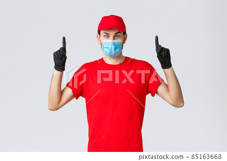 Covid-19, self-quarantine, online shopping and shipping concept. Surprised delivery guy in red uniform and cap, raising eyebrows amazed or excited, showing promo top, pointing up, wear medical mask 85163668