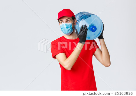 Takeaway, food and groceries delivery, covid-19 contactless orders concept. Carefree courier in red uniform cap, face mask and gloves, hold bottled water on shoulder, look away with smile 85163956