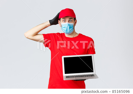 Customer support, covid-19 delivery packages, online orders processing concept. Confused and worried courier in red uniform, face mask and gloves, look nervous, show laptop screen 85165096