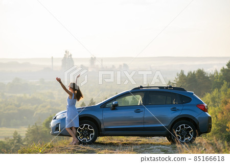 Young female driver resting near her car raising up her hands on warm summer evening. Travel destinations concept. 85166618