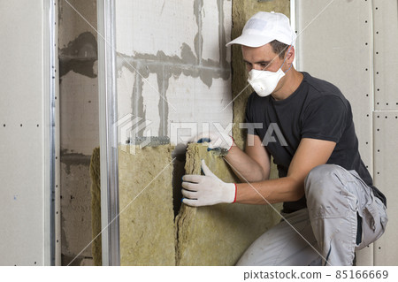 Worker in protective respirator mask insulating rock wool insulation in wooden frame for future house walls for cold barrier. Comfortable warm home, economy, construction and renovation concept Worker in protective respirator mask insulating rock wool insulation in wooden frame for future house walls for cold barrier. Comfortable warm home, economy, construction and renovation concept 85166669