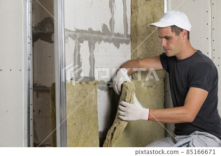 Worker in protective gloves insulating rock wool insulation in wooden frame for future house walls for cold barrier. Comfortable warm home, economy, construction and renovation concept Worker in protective gloves insulating rock wool insulation in wooden frame for future house walls for cold barrier. Comfortable warm home, economy, construction and renovation concept 85166671