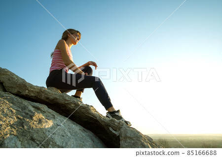 Woman hiker sitting on a steep big rock enjoying warm summer day. Young female climber resting during sports activity in nature. Active recreation in nature concept. 85166688