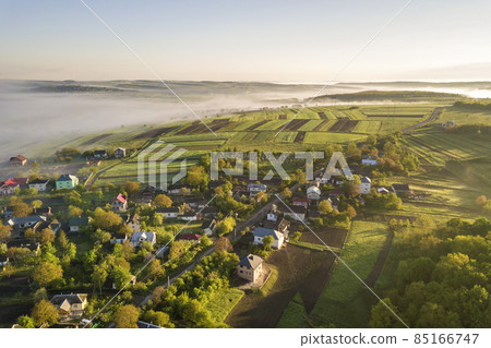 View from above of white fog on village house roofs among green trees under bright blue sky. Spring misty landscape panorama at dawn. 85166747