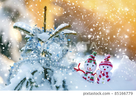 Two small funny toys baby snowman in knitted hats and scarves in deep snow outdoors near pine tree branch. Happy New Year and Merry Christmas greeting card. 85166779