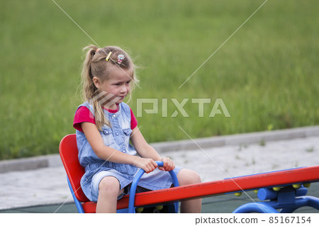 Small cute young blond child girl sits on see- saw swing on warm sunny day on bright green blurred background. Joys and sorrows of childhood, outdoor activity concept. 85167154